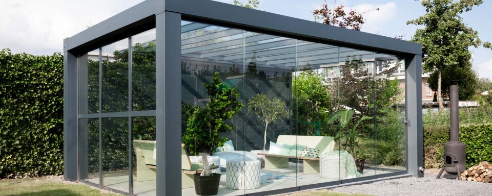 Vrijstaande overkapping met steellook schuifwand, volledig uitgevoerd in glas en ingericht als groene loungeplek in de tuin.