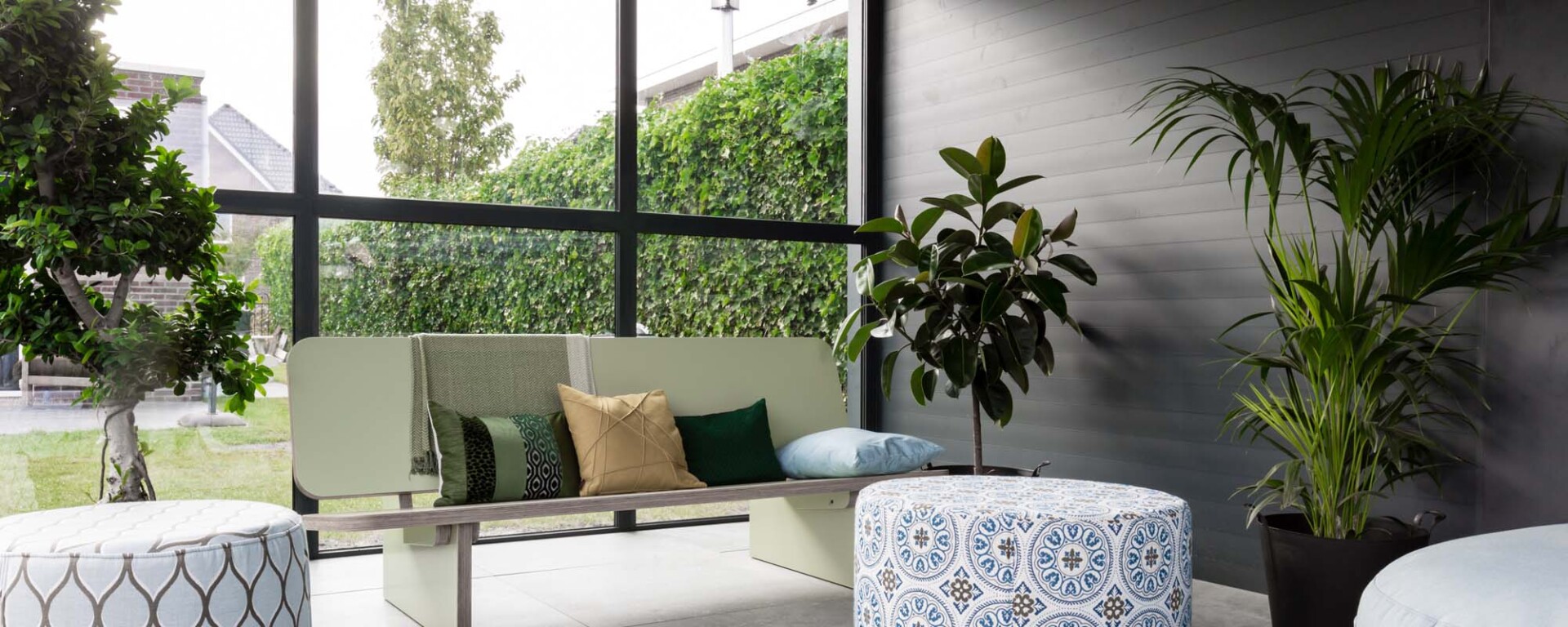 Stijlvolle zithoek achter schuifpui met zwart profiel, geplaatst in een moderne overkapping met uitzicht op een groene tuin.
