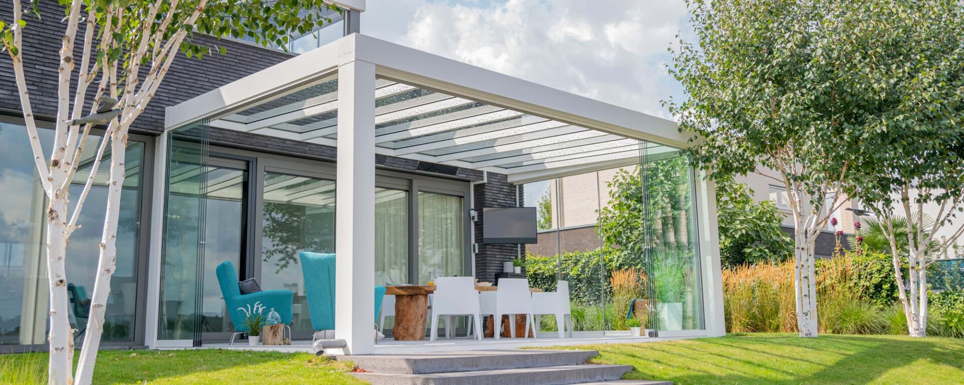 Witte Verasol Cube veranda met glazen wanden en dak, modern ingericht als lounge- en eetruimte in een groene tuin.