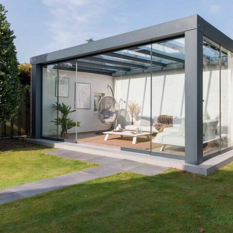 Vrijstaande Verasol Cube veranda met glazen schuifwanden, modern ingericht met loungebank en hangstoel in een groene tuin.